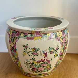 Könnte beinhalten: Ein runder, cremefarbener Keramik-Blumentopf mit breitem Rand. Der Topf ist mit bunten Blumen- und Vogelmotiven in Rosa, Gelb und Grün verziert. Der Rand hat einen roten Rand. Der Topf steht auf einer Holzoberfläche.