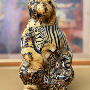 May include: A ceramic bear figurine with a unique design. The bear's body is adorned with patterns of zebra stripes, leopard spots, and tiger stripes, in shades of brown, black, and tan. The bear is in a seated position.
