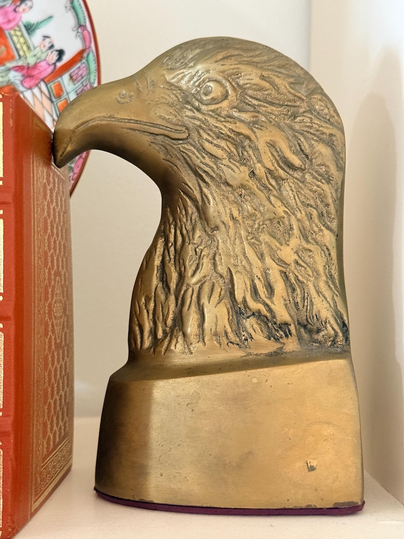 May include: A brass eagle head sculpture, likely a bookend, with detailed feather texture and a strong beak. The sculpture has a rectangular base and is set against a neutral background. The eagle's head is angled slightly upward.