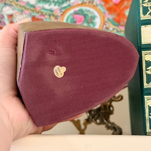 May include: A burgundy bookend with a gold-colored top and a label that reads "Made in Korea". The bookend has a soft, velvety texture. The background includes a decorative plate and a green book.