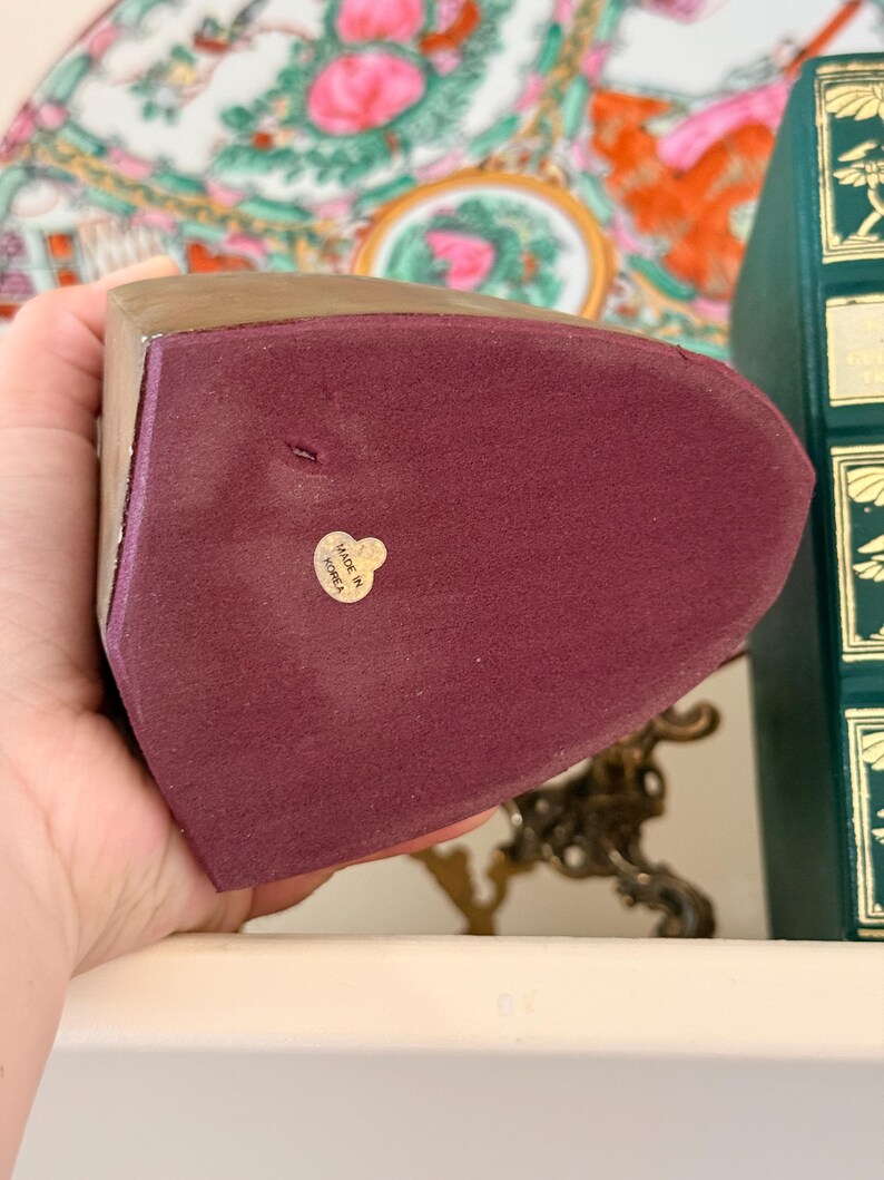 May include: A burgundy bookend with a gold-colored top and a label that reads "Made in Korea". The bookend has a soft, velvety texture. The background includes a decorative plate and a green book.