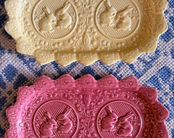 May include: Two ceramic serving trays, one pale yellow and one pink, each with a scalloped edge and a central design of two rabbits. The trays have a decorative, embossed floral pattern and are set against a blue and white patterned background.