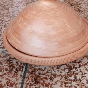 May include: A terracotta tagine pot with a conical lid and a wide, shallow base. The pot has a textured, ridged surface and is designed for slow cooking. The tagine is a traditional North African cooking vessel.