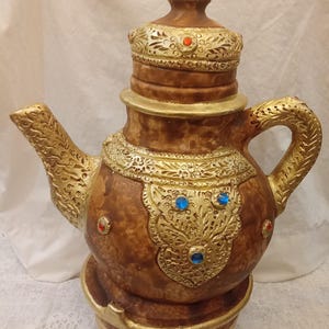 Könnte beinhalten: Eine dekorative braune Teekanne mit goldenen Akzenten und aufwendigen Details. Die Teekanne hat einen Deckel, einen Griff und einen Ausguss, verziert mit goldenem Filigran und roten und blauen juwelenartigen Verzierungen. Der Sockel ist ebenfalls goldfarben.