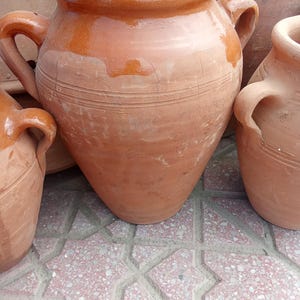 May include: Three terracotta pots with handles. The pots are brown and have a rough texture. The pots are sitting on a tiled floor.
