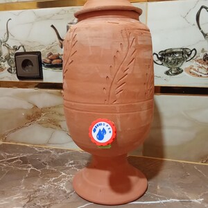 May include: A terracotta water dispenser with a lid and a base. The dispenser has carved leaf designs and a blue and white "Open" sticker near the spigot. The dispenser is on a countertop with a patterned tile backsplash.