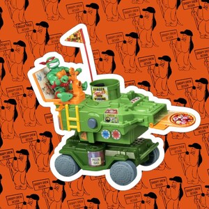 May include: A green Teenage Mutant Ninja Turtles pizza-themed vehicle toy featuring a Raphael figure, a red flag with a pizza slice graphic, and stickers labeled DANGER TURTLES AT WORK, CHEESE, and PIZZA GAS, set against an orange background.