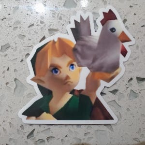 May include: A sticker featuring a cartoon character with blonde hair, blue eyes, and pointed ears, holding a chicken. The character is wearing a green tunic and a black hat. The chicken is gray with a red comb and beak.