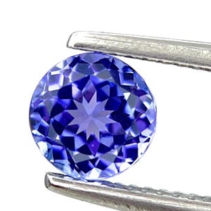 Puede incluir: Una gema redonda, facetada, de color azul intenso, es sostenida por unas pinzas plateadas sobre un fondo blanco. Las facetas de la gema crean un patrón de estrella en el centro. La gema tiene aproximadamente 1,3 cm de diámetro.