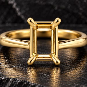 Könnte beinhalten: Ein Ring aus poliertem Gelbgold mit einer rechteckigen Fassung und vier Krappen, die für einen Edelstein ausgelegt sind. Die Ringschiene ist glatt und abgerundet und verbindet sich mit dem leeren, rechteckigen Rahmen in der Mitte.