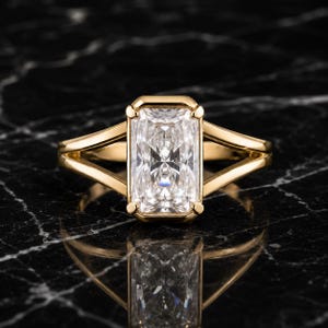 Peut inclure: Une bague en or avec une grande pierre précieuse transparente de taille radiante rectangulaire maintenue par quatre griffes. L'anneau est en or jaune poli et se divise en deux branches distinctes de chaque côté, créant un design géométrique ouvert.