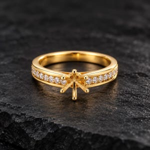 Puede incluir: Un anillo de oro con un engaste central de seis puntas diseñado para sostener una piedra preciosa. La banda está acentuada con una hilera de pequeñas piedras claras de corte redondo incrustadas a los lados, creando un efecto brillante.