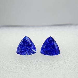 Puede incluir: Dos gemas triangulares, facetadas, de color azul intenso. Las gemas se exhiben sobre una superficie blanca, mostrando su corte y color brillantes. Estas gemas son probablemente para la fabricación de joyas.