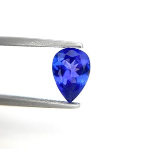 Puede incluir: Una gema azul oscuro en forma de pera, sujeta con pinzas plateadas. La gema está facetada, mostrando su estructura interna y color. El fondo es blanco liso, resaltando el tono vibrante de la gema.