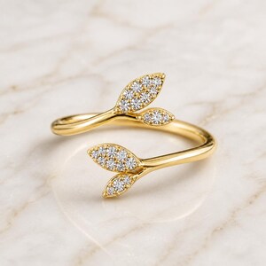 Puede incluir: Un anillo abierto de oro con dos motivos en forma de hoja en cada extremo. Cada hoja está incrustada con pequeñas piedras redondas y transparentes que captan la luz. La banda es lisa y pulida, creando un diseño abierto y envolvente.