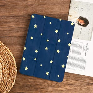 May include: A navy blue tablet case with a pattern of yellow stars. The case is rectangular and has a folding cover. The background is a wooden surface with a woven basket and a magazine.