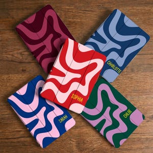 Op de afbeelding: Vijf rechthoekige tablethoezen met golvende, meerkleurige patronen. De hoezen zijn uitgevoerd in bordeauxrood, licht- en donkerblauw, rood en roze, blauw en roze, en groen en paars. Drie hoezen zijn voorzien van de namen Charlotte, Sophia en Chloe.