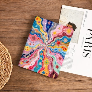 May include: A colorful journal with a vibrant, abstract design featuring swirling lines and stars in shades of pink, blue, yellow, and orange. The journal is open, revealing a blank page. A magazine with the word "PARIS" is next to the journal.