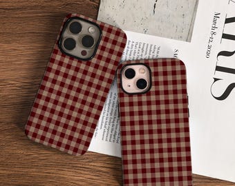 May include: Two phone cases with a red and tan gingham pattern. The cases have a black camera lens frame. One case is angled towards the viewer, while the other is upright. The background includes a wooden surface and a magazine.