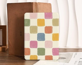 May include: A cream-colored tablet case with a colorful checkerboard pattern of squares in pink, green, yellow, orange, and blue. The case is propped up next to a brown book on a wooden surface.