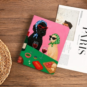 Może przedstawiać: Różowy notatnik z ilustracjami dwóch psów w okularach przeciwsłonecznych i chustach na głowie, delektujących się winem i przekąskami. Słowo "SASSY" znajduje się na czerwonym pudełku. W tle magazyn ze słowem "PARIS".