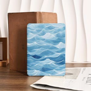Puede incluir: Una funda rectangular para tableta con un diseño de olas azules y blancas. La funda está colocada sobre una superficie de madera, junto a un libro antiguo y un periódico. El diseño de olas es un patrón repetitivo de olas oceánicas estilizadas.