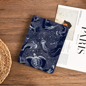 May include: A dark blue notebook cover featuring a white illustration of various sea creatures, including whales, jellyfish, and a sea turtle, set against a starry background. The cover is lying on a wooden surface next to a magazine.