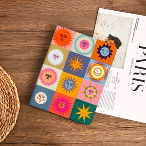 Op de afbeelding: Een kleurrijk notitieboek met een raster van zonne-illustraties in verschillende stijlen en kleuren. De omslag bevat oranje, gele, roze, blauwe en groene zonnen met lachende gezichten. Een tijdschrift met het woord "PARIS" is gedeeltelijk zichtbaar.