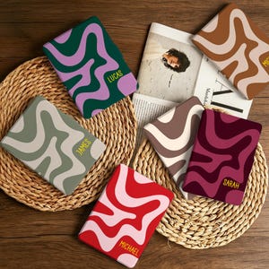 May include: Six rectangular e-reader covers featuring wavy, abstract patterns in various color combinations including green and purple, sage and cream, red and pink, brown and tan, and maroon and dark pink, with personalized names printed in yellow.
