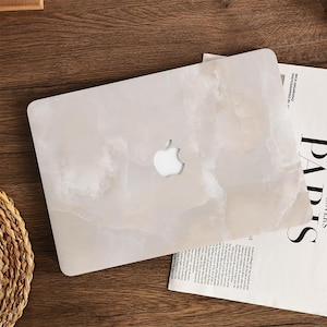 May include: A white laptop case with a marble effect, featuring the Apple logo cutout. The case is on a wooden surface next to a magazine. The marble pattern has shades of white and beige.
