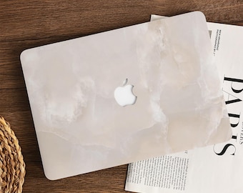 Peut inclure: Coque d'ordinateur portable blanche avec un motif marbré, avec le logo Apple découpé. Le boîtier repose sur une surface en bois à côté d'un magazine. Le motif marbré présente des nuances de blanc et de beige.