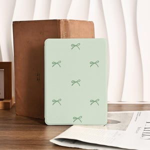 Peut inclure: Une coque de tablette vert clair ornée d'un motif de petits nœuds verts. Elle est posée à côté d'un livre marron vintage. L'arrière-plan comprend une surface en bois et un journal.
