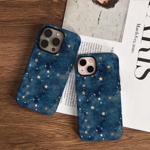 May include: Two smartphone cases with a dark blue watercolour design featuring gold stars and white speckles. The cases have a glossy finish and are on a wooden surface. The cases are for a phone with multiple cameras.