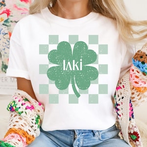 May include: White t-shirt with a green four-leaf clover graphic on a checkered background. The clover has the word "laki" written inside.