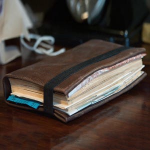 May include: A brown leather journal with a black elastic band closure. The journal is filled with pages and has a blue tag sticking out. The journal is on a dark wooden surface.