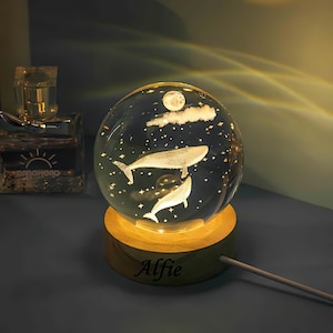 Könnte beinhalten: Eine Nachtlampe aus klarem Glas in Kugelform mit einem Holzsockel. Die Kugel zeigt einen weißen Wal und einen kleineren Wal, die unter einem Halbmond und Wolken schwimmen. Der Sockel ist mit dem Namen "Alfie" graviert.
