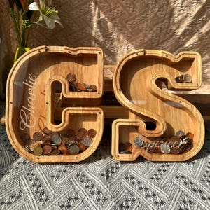 Può includere: Due salvadanai in legno a forma di lettera, una "G" e una "S", pieni di monete. La "G" ha inciso "Charlie", e la "S" ha "Spencer". I salvadanai sono in legno chiaro e hanno frontali in acrilico trasparente.