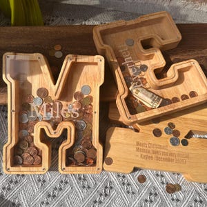 May include: Wooden letter-shaped and key-shaped coin banks filled with coins and a dollar bill. The letter "M" is engraved with the name "Miles". The key-shaped bank has the name "Everhetti" engraved on it. The bottom of the key-shaped bank has a message.
