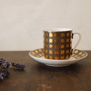 Puede incluir: Un juego de taza y platillo vintage con un patrón geométrico marrón y dorado. La taza y el platillo son blancos con un patrón de puntos marrones y dorados. Un pequeño manojo de lavanda seca está sobre la mesa.