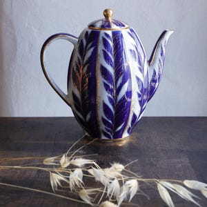 May include: A vintage porcelain teapot with a blue and white leaf pattern. The teapot has a gold handle, spout, and lid finial. It sits on a dark wooden surface with dried flowers in the foreground.