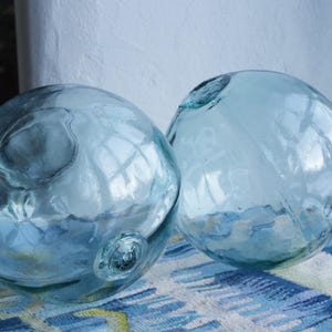 Puede incluir: Dos flotadores de vidrio esféricos transparentes con un tinte azul claro. Los flotadores tienen una superficie texturizada y una pequeña abertura en la parte superior. Están sobre una tela estampada azul y blanca.
