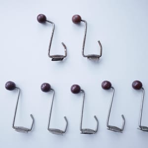 Pode incluir: Sete ganchos de metal vintage com um botão esférico castanho escuro no topo. Os ganchos têm um design de metal prateado curvo com uma pequena base retangular. Os ganchos estão dispostos sobre um fundo branco.