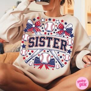 May include: A beige sweatshirt with a graphic design featuring two crossed baseball bats, baseballs, stars, and a red, white, and blue bow. The text "SISTER" is printed in black letters on the graphic.