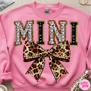 Pu&ograve; includere: Felpa rosa con la scritta "MINI" in oro, nero e strass. Un fiocco leopardato con strass &egrave; centrato sotto la scritta. Una borraccia con stampa leopardata &egrave; visibile in basso a destra.