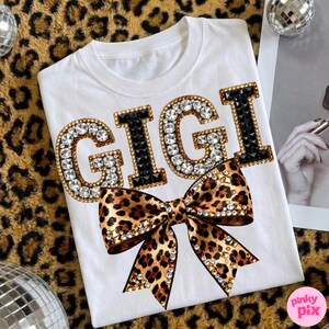 May include: White t-shirt with the name "GIGI" in large, sparkling letters. The letters "G" are outlined in gold and filled with clear rhinestones, while the "I"s are black. A leopard print bow with rhinestone accents is below the name.