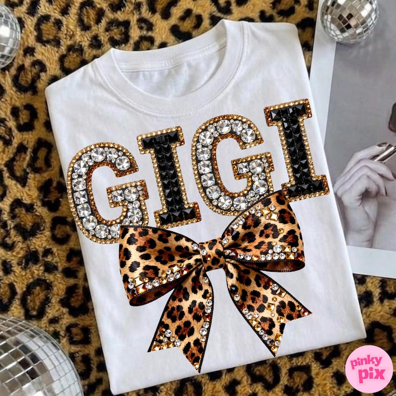 May include: White t-shirt with the name "GIGI" in large, sparkling letters. The letters "G" are outlined in gold and filled with clear rhinestones, while the "I"s are black. A leopard print bow with rhinestone accents is below the name.