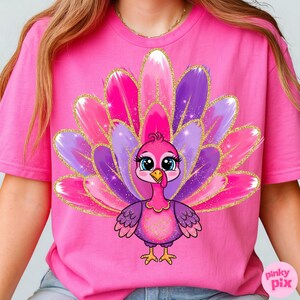 May include: Pink t-shirt featuring a cartoon turkey with a vibrant, colorful tail. The turkey has a pink body, purple wings, and a yellow beak. The tail feathers are pink, purple, and white with gold glitter accents. The text "pinky pix" is in the bottom right corner.
