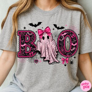 Puede incluir: Camiseta gris con un dise&ntilde;o de Halloween rosa y negro. La palabra "BOO" est&aacute; escrita con letras rosas, un fantasma, murci&eacute;lagos y telara&ntilde;as. El fantasma tiene un lazo rosa y las letras est&aacute;n delineadas con encaje negro.