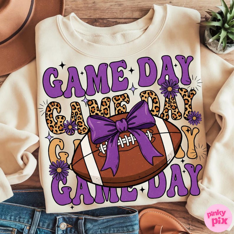 May include: Cream-colored sweatshirt with the text "GAME DAY" in purple, leopard print, and gold lettering. A brown football with a purple bow is centered on the shirt. Purple flowers and stars accent the design.