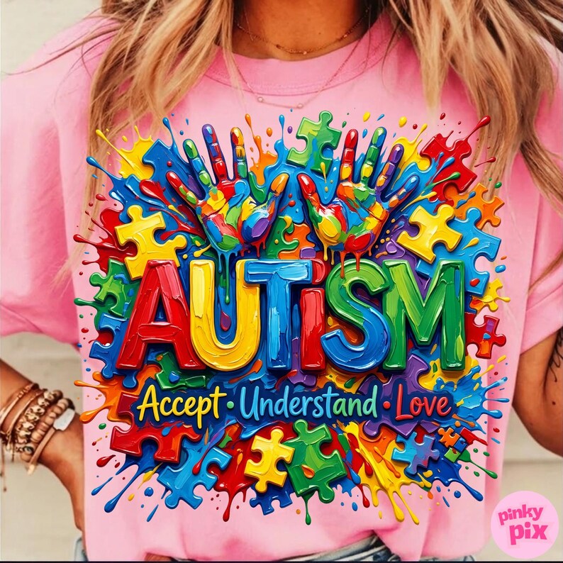 Op de afbeelding: Roze T-shirt met een kleurrijk grafisch ontwerp. Het ontwerp toont het woord "AUTISME" in grote, veelkleurige letters, omringd door puzzelstukjes en handafdrukken. De woorden "Accepteer, Begrijp, Liefde" staan eronder.